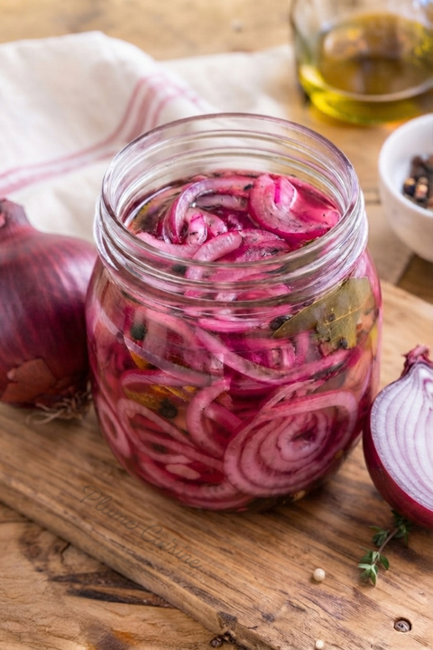 Bocal en verre rempli d’oignons rouges marinés roses, avec quelques épices, posé sur une planche en bois.